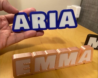 Könnte beinhalten: 3D-gedruckte Namensschilder. Ein Schild schreibt "ARIA" in weißen Buchstaben mit blauem Rand, gehalten von einer Hand. Ein weiteres Schild schreibt "EMMA" in weißen Buchstaben mit roségoldenem Rand. Ein drittes Schild mit schwarzem Rand ist teilweise sichtbar.