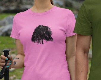 Op de afbeelding: Een vrouw draagt een roze T-shirt met een zwart silhouet van een treurwilg op de voorkant. Ze draagt een zonnebril en heeft een tatoeage op haar arm. Ze houdt een wandelstok vast.