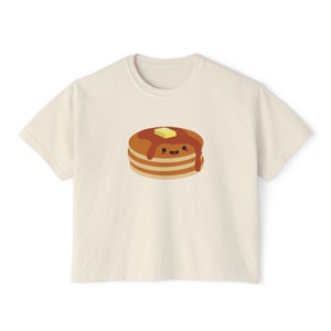 Puede incluir: Camiseta de color crema con una pila de dibujos animados de panqueques con jarabe y una nuez de mantequilla. Los panqueques tienen una cara sonriente. La camiseta tiene mangas cortas y cuello redondo.