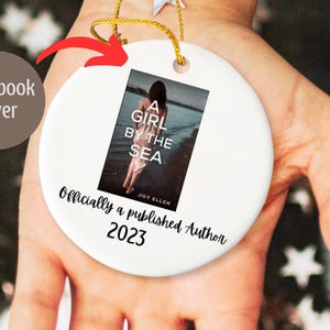 Könnte beinhalten: Weißer Keramik-Anhänger mit einem Buchcover-Bild und dem Text "A Girl by the Sea" von Joy Ellen. Der Anhänger enthält auch den Text "Officially a published Author 2023".