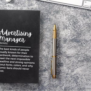 May include: Black leather journal with the text "Advertising Manager /noun/ The best kinds of people usually known for their hard work, determination to meet the next impossible deadline and strong opinions about fonts, colours, and why Comic Sans should never exist."