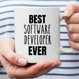 May include: White ceramic mug with the text "BEST SOFTWARE DEVELOPER EVER" in black, held by a person. The mug has a handle and is set against a striped background.