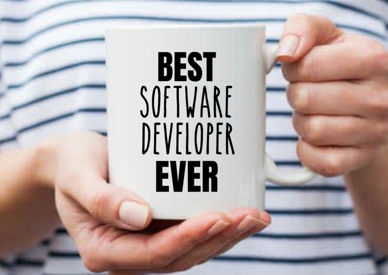 May include: White ceramic mug with the text "BEST SOFTWARE DEVELOPER EVER" in black, held by a person. The mug has a handle and is set against a striped background.