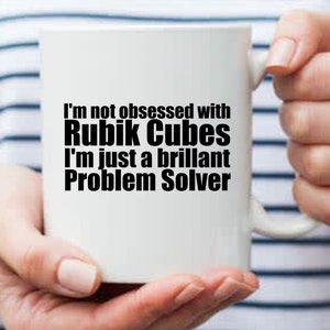 Può includere: Una tazza in ceramica bianca con testo nero che recita "Non sono ossessionato dai Cubi di Rubik, sono solo un brillante risolutore di problemi"