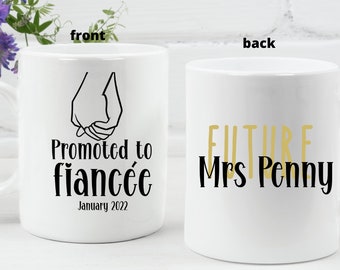 May include: White ceramic mug with black text that reads "Promoted to Fiancée January 2022" and a black line drawing of two hands holding each other. The back of the mug has gold text that reads "Future Mrs. Penny".