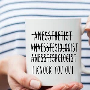 May include: White ceramic coffee mug with black text that reads "Anesthetist" with a line through it, "Anaesthetist" with a line through it, "Anaesthetist" with a line through it, and "I knock you out".