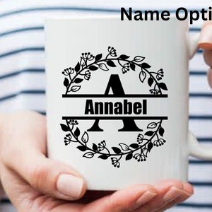 May include: White ceramic mug with a black floral wreath design, the letter "A", and the name "Annabell" in a bold, black font. The text "Name Option 2" is at the top. The mug is held by a person.