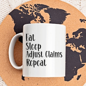 Könnte beinhalten: Eine weiße Keramiktasse mit schwarzem Text, der "Eat Sleep Adjust Claims Repeat" lautet. Die Tasse steht auf einem Korkuntersetzer mit einem schwarzen Weltkartenmotiv.