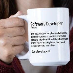 May include: White ceramic mug with the text "Software Developer" and a definition. The definition describes software developers as people known for their hard work and fast typing skills. The mug is held by a person.