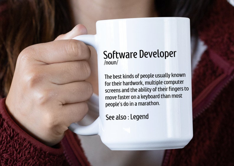 May include: White ceramic mug with the text "Software Developer" and a definition. The definition describes software developers as people known for their hard work and fast typing skills. The mug is held by a person.