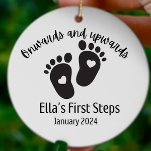 Op de afbeelding: Witte keramische ornament met zwarte voetafdrukken en harten, de tekst "Onwards and upwards Ella's First Steps January 2024" is gedrukt op de ornament.