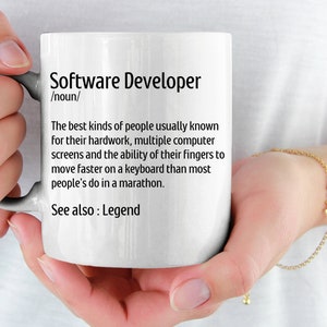 May include: White ceramic mug with the text "Software Developer" and a definition. The definition describes the skills and traits of software developers. The mug is held by a person.