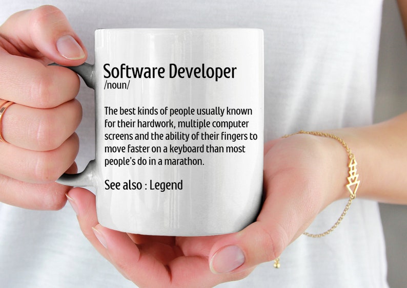 May include: White ceramic mug with the text "Software Developer" and a definition. The definition describes the skills and traits of software developers. The mug is held by a person.