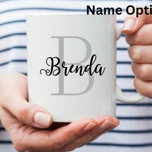 May include: White ceramic mug with the name "Brenda" in a stylish script font, superimposed over a large gray letter "B". The mug is held by a person with a striped background and the text "Name Option 1" at the top.