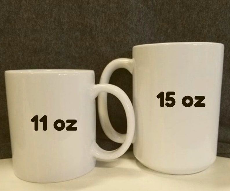 May include: Two white ceramic mugs with curved handles are displayed. The smaller mug is labeled "11 oz" and the larger mug is labeled "15 oz". Both mugs are plain white and have a glossy finish.