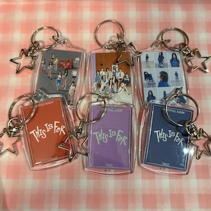 Pode incluir: Chaveiros com imagens de um grupo musical, com um pingente em forma de estrela. Os chaveiros têm uma moldura de plástico transparente com um porta-chaves prateado. Os chaveiros têm um fundo vermelho, roxo e azul com o texto "THIS IS FOR".