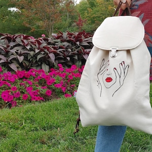 May include: A cream-coloured canvas backpack featuring a minimalist black line art illustration of a face with hands covering the eyes and bright red lips. The bag has a fold-over flap with a silver metal clasp and brown fabric shoulder straps.