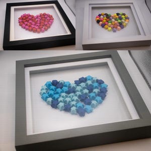 Puede incluir: Tres decoraciones de corazón de origami enmarcadas. El primer marco es negro y presenta un corazón rosa. El segundo marco es blanco y presenta un corazón multicolor. El tercer marco es gris y presenta un corazón azul y azul claro.