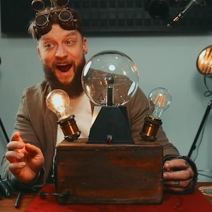May include: A man wearing goggles and a beard is holding a wooden box with a clear glass sphere, two light bulbs, and a black base. The box is sitting on a red table.