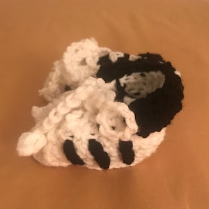May include: Handmade crochet baby booties in a panda bear design. The booties are primarily white with black accents, including the top of the shoe and stripes. The soft, textured yarn creates a cozy and adorable look for newborns.