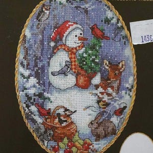 May include: An oval counted cross stitch ornament kit featuring a snowman wearing a red hat and plaid scarf, surrounded by a deer, rabbit, birds, and a basket of berries. The design is framed in a gold-toned border with ornate metal accents.