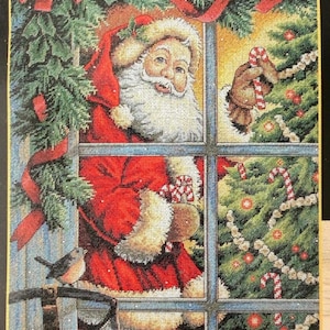 May include: A cross-stitch embroidery depicting Santa Claus in a red coat with white fur trim, looking through a window. He holds a candy cane near a decorated Christmas tree. A small bird sits on a sled frame in the foreground.