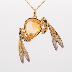 Op de afbeelding: Een gouden ketting met een hanger met een grote, gefacetteerde, oranje edelsteen omringd door twee gouden wespen met zwarte en blauwe emaildetails.