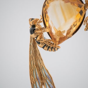 Op de afbeelding: Een gouden ketting met een hanger met een grote, gefacetteerde topaas-edelsteen omringd door twee gedetailleerde, gouden wespen met zwarte en witte accenten. De wespen hebben doorschijnende, blauwe en groene vleugels.