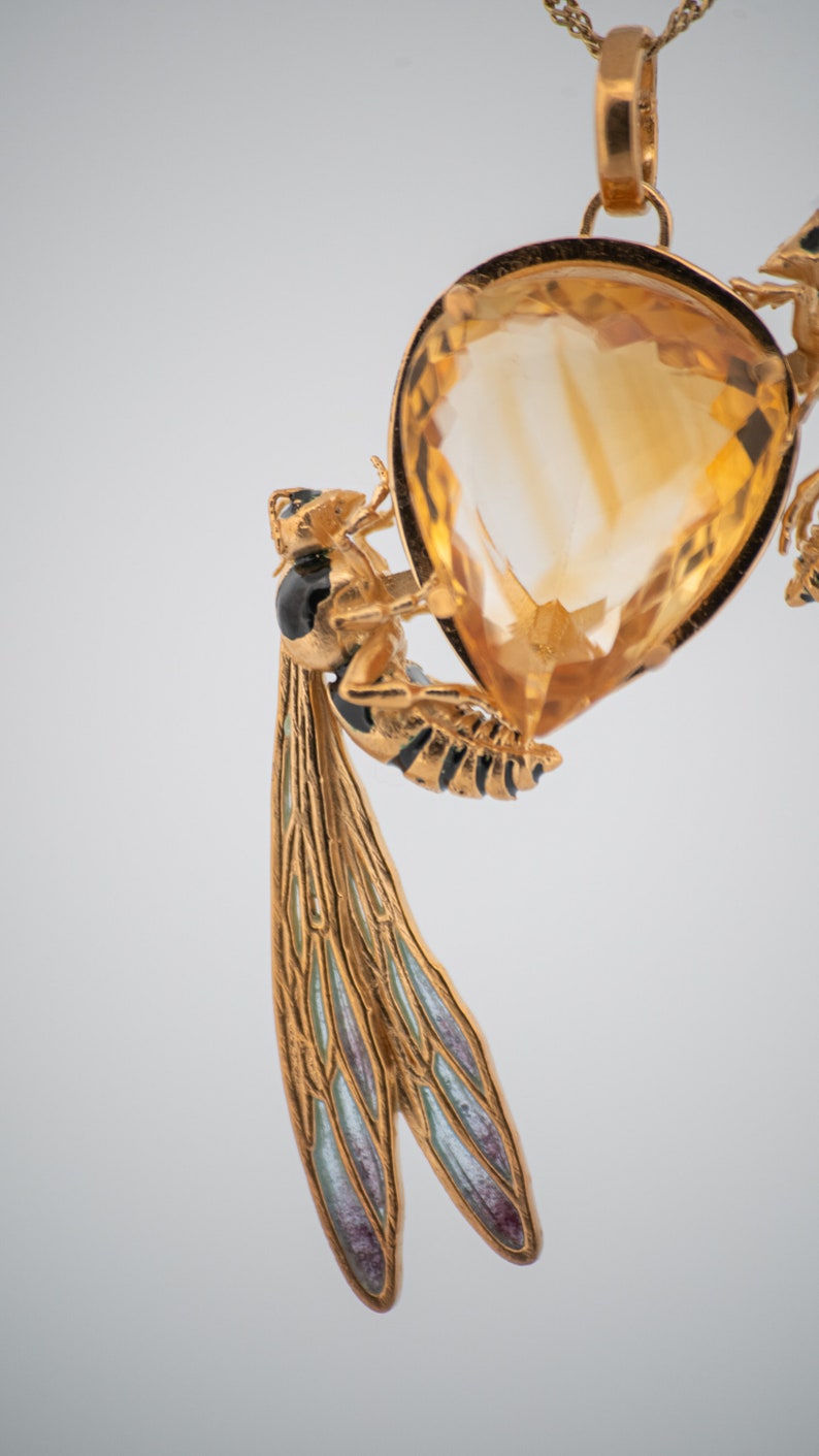 Op de afbeelding: Een gouden ketting met een hanger met een grote, gefacetteerde topaas-edelsteen omringd door twee gedetailleerde, gouden wespen met zwarte en witte accenten. De wespen hebben doorschijnende, blauwe en groene vleugels.