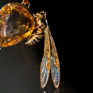 Op de afbeelding: Een gouden ketting met een hanger, een grote, gefacetteerde, gele edelsteen en een gedetailleerde, gouden wesp met blauwe emaille vleugels.
