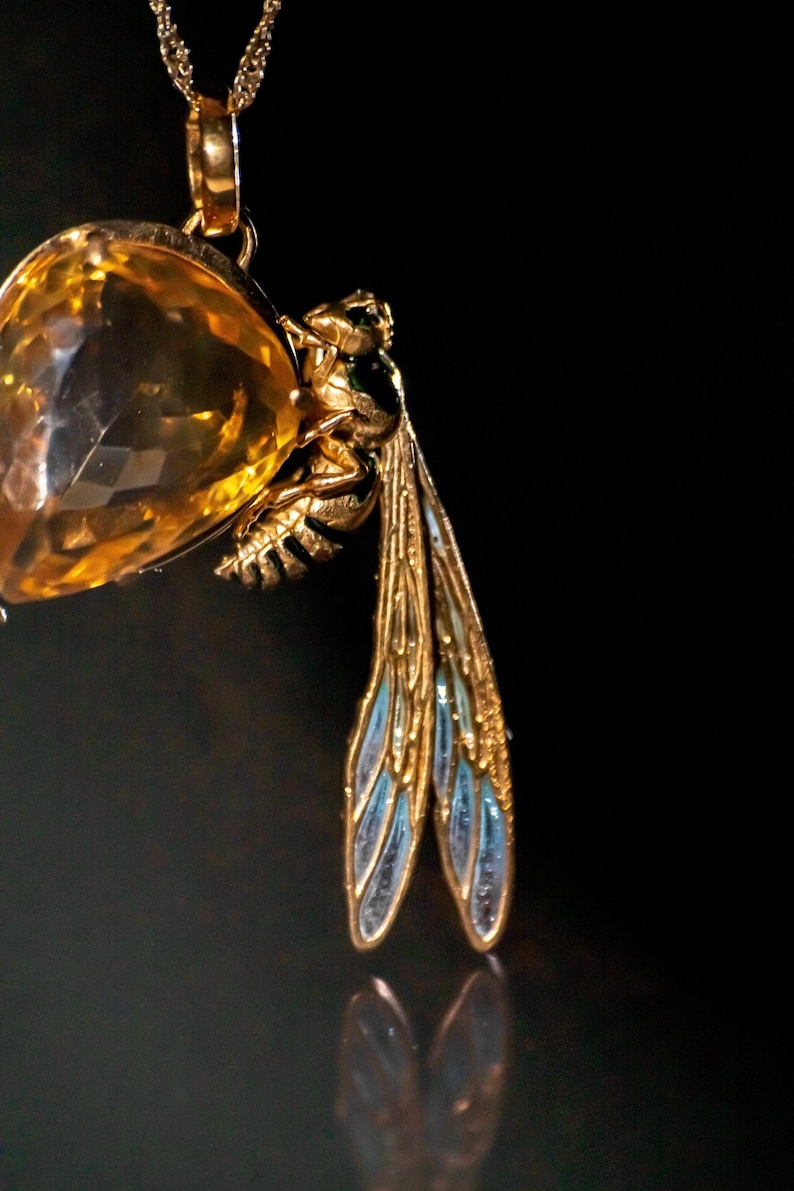 Op de afbeelding: Een gouden ketting met een hanger, een grote, gefacetteerde, gele edelsteen en een gedetailleerde, gouden wesp met blauwe emaille vleugels.