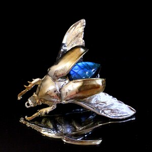 Puede incluir: Una escultura de escarabajo de plata y oro con alas azules iridiscentes. Las alas están abiertas y el escarabajo está en vuelo.