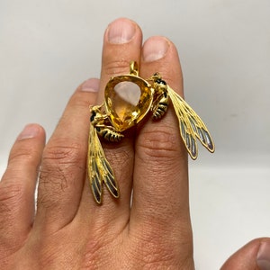 Op de afbeelding: Een gouden hanger met twee wespen met zwarte en gele markeringen, die een grote, gefacetteerde, gele edelsteen vasthouden.