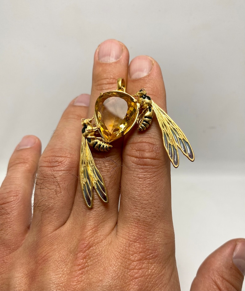 Op de afbeelding: Een gouden hanger met twee wespen met zwarte en gele markeringen, die een grote, gefacetteerde, gele edelsteen vasthouden.