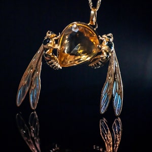 Op de afbeelding: Een gouden ketting met een hanger met een grote, gefacetteerde, amberkleurige edelsteen. Aan de zijkanten van de edelsteen zijn twee gouden bijfiguren met zwarte en blauwe emaille vleugels bevestigd.