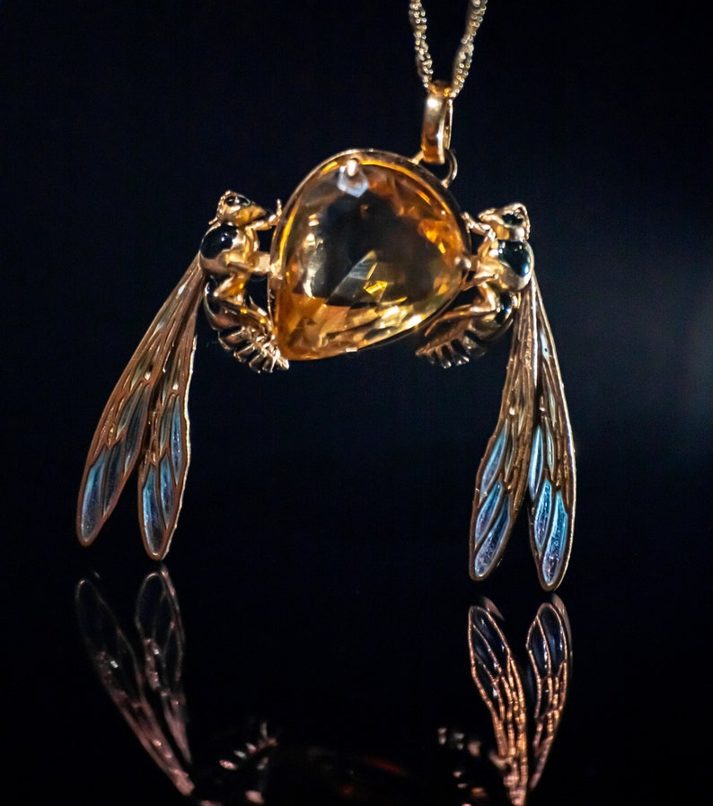 Op de afbeelding: Een gouden ketting met een hanger met een grote, gefacetteerde, amberkleurige edelsteen. Aan de zijkanten van de edelsteen zijn twee gouden bijfiguren met zwarte en blauwe emaille vleugels bevestigd.