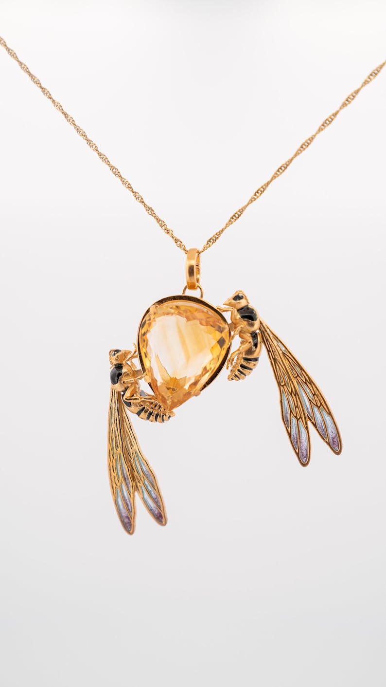 Op de afbeelding: Een gouden ketting met een hanger met een grote, gefacetteerde, oranje edelsteen omringd door twee gouden wespen met zwarte en blauwe emaildetails.