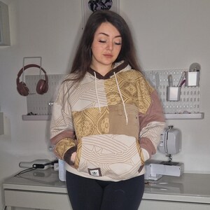 May include: A beige and brown patchwork hoodie with a kangaroo pocket and a drawstring hood. The hoodie features a paisley pattern and a small logo on the front. The sleeves are a solid mauve colour. The person is wearing black leggings.