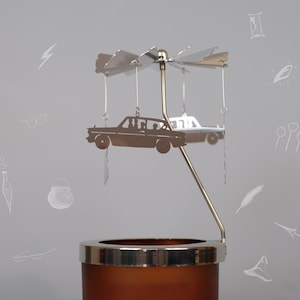 May include: A silver metal carousel candle holder featuring a silhouette of a car and passengers. The carousel sits on a brown candle holder. The background is grey with white drawings of various Harry Potter themed objects.