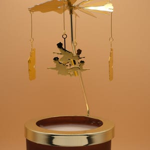 May include: A gold-colored candle carousel with a brown base and a gold rim. The carousel features a spinning top with cut-out shapes and hanging gold ornaments. The candle carousel is designed to rotate when the candles are lit.