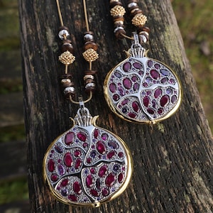 May include: Two necklaces featuring pomegranate pendants with gold-coloured borders and silver-coloured centres. The pendants are adorned with red and purple gemstones. The necklaces hang on thin, gold cords with decorative beads.
