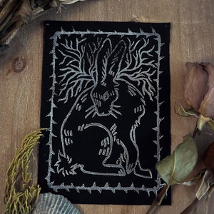May include: A black fabric patch with a silver-colored design of a rabbit with a tree growing out of its head. The design is surrounded by a border of thorns.