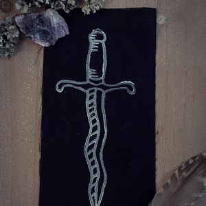 May include: A white embroidered dagger design on a black fabric patch. The patch is surrounded by natural elements like lichen and feathers.