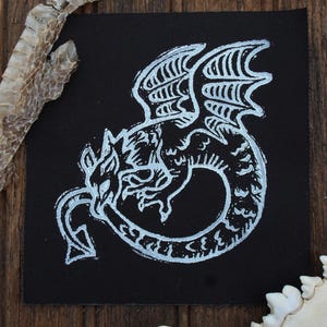 May include: A white dragon illustration on a black square. The dragon is depicted with wings and a long tail. The image is surrounded by a snake skin and a bone on a wooden surface.