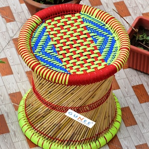 May include: A woven bamboo stool with a red, yellow, and blue woven top. The stool has a red and yellow woven band around the base. The stool has a label that says "GUNEER" on the side.