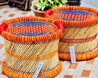 Peut inclure: Deux tabourets ronds en bambou tressé avec des accents de corde tressée orange, rouge et bleu. Les tabourets portent une étiquette indiquant "Gunee".