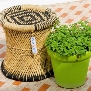 Op de afbeelding: Een geweven bamboe kruk met zwarte bies zit op een geblokte tegelvloer naast een groene pot gevuld met een weelderige groene plant. De kruk heeft een label met de tekst "Gunee".