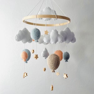 Op de afbeelding: Een houten babymobiel met gehaakte ballonnen in blauw, beige en perzikkleuren, witte viltwolken en houten sterren.