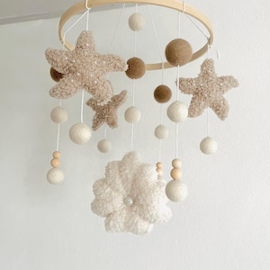 Könnte beinhalten: Baby-Mobile mit Holzring, beigen Plüschsternen und Filzkugeln in Weiß und Braun. Eine weiße Blume und Holzperlen sind ebenfalls aufgehängt. Das Mobile hängt an weißer Schnur.
