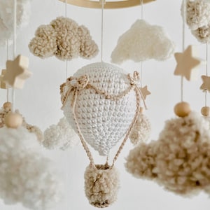 Könnte beinhalten: Handgefertigtes Babymobile mit einem weißen gehäkelten Heißluftballon und einem beigen Korb, umgeben von flauschigen Wolken und Holzstern-Akzenten. Das Mobile hängt an einem Holzreifen und schafft eine skurrile Kinderzimmerdekoration.
